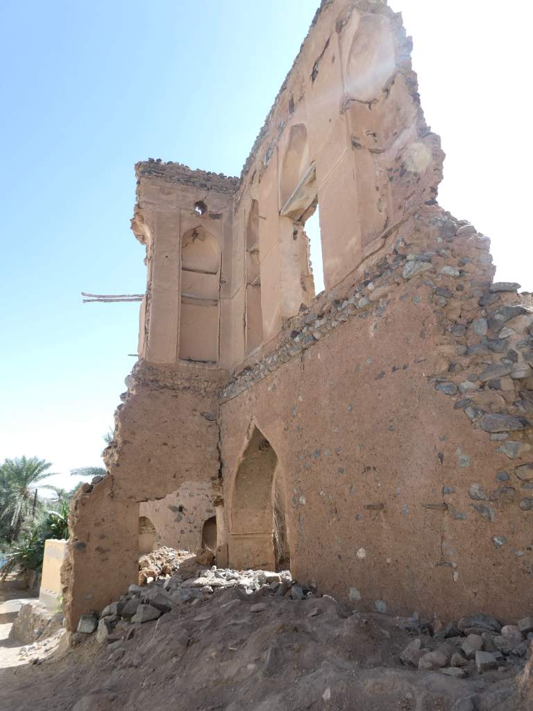 Ruines d'une ancienne structure en terre, avec des murs effondrés et des fenêtres ouvertes, devant un ciel bleu clair.