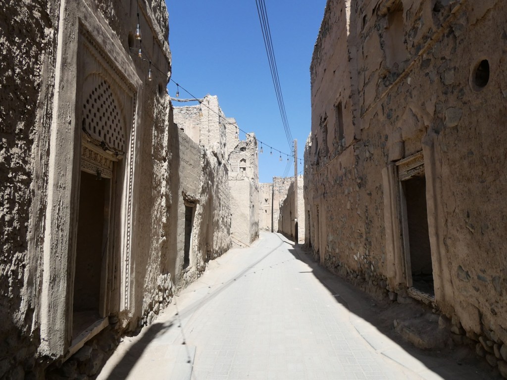 Une ruelle étroite dans un village ancien avec des murs en pierre et en terre, baignée de lumière naturelle sous un ciel bleu.