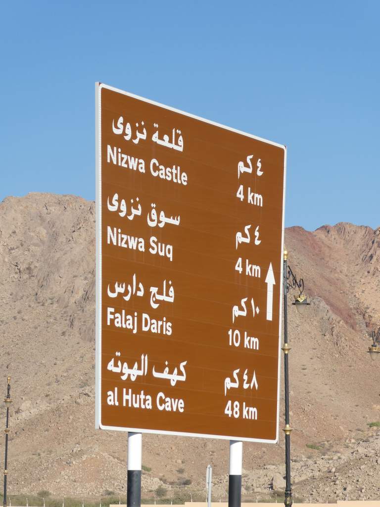 Panneau de signalisation indiquant les distances vers le Château de Nizwa, le Souk de Nizwa, le Falaj Daris et la grotte al Huta.