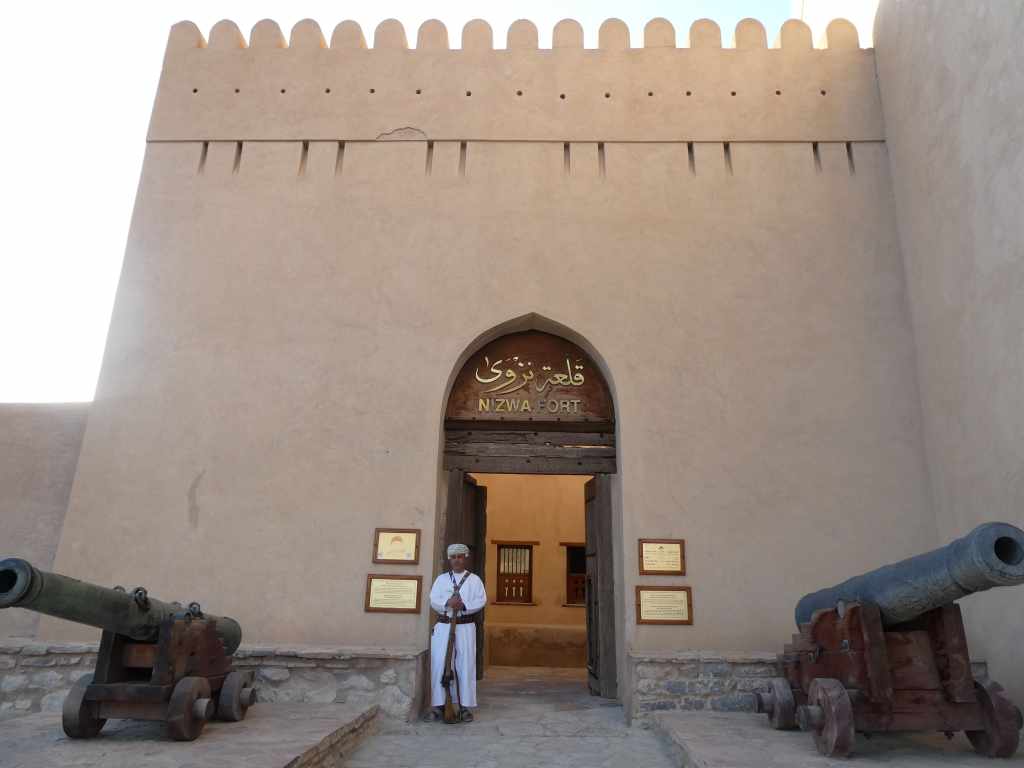 Fort de Nizwa avec sa porte d'entrée et des canons en bois de chaque côté, sous un ciel clair.