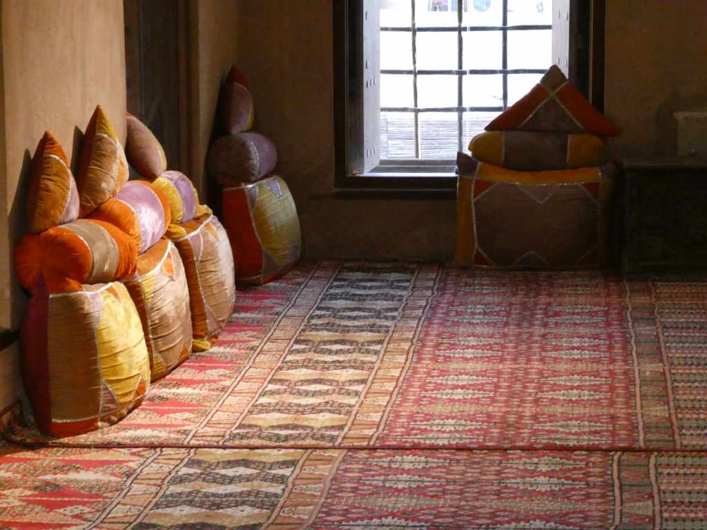 Intérieur d'une pièce avec des coussins colorés empilés contre le mur et un tapis décoratif au sol, éclairé par la lumière naturelle d'une fenêtre.