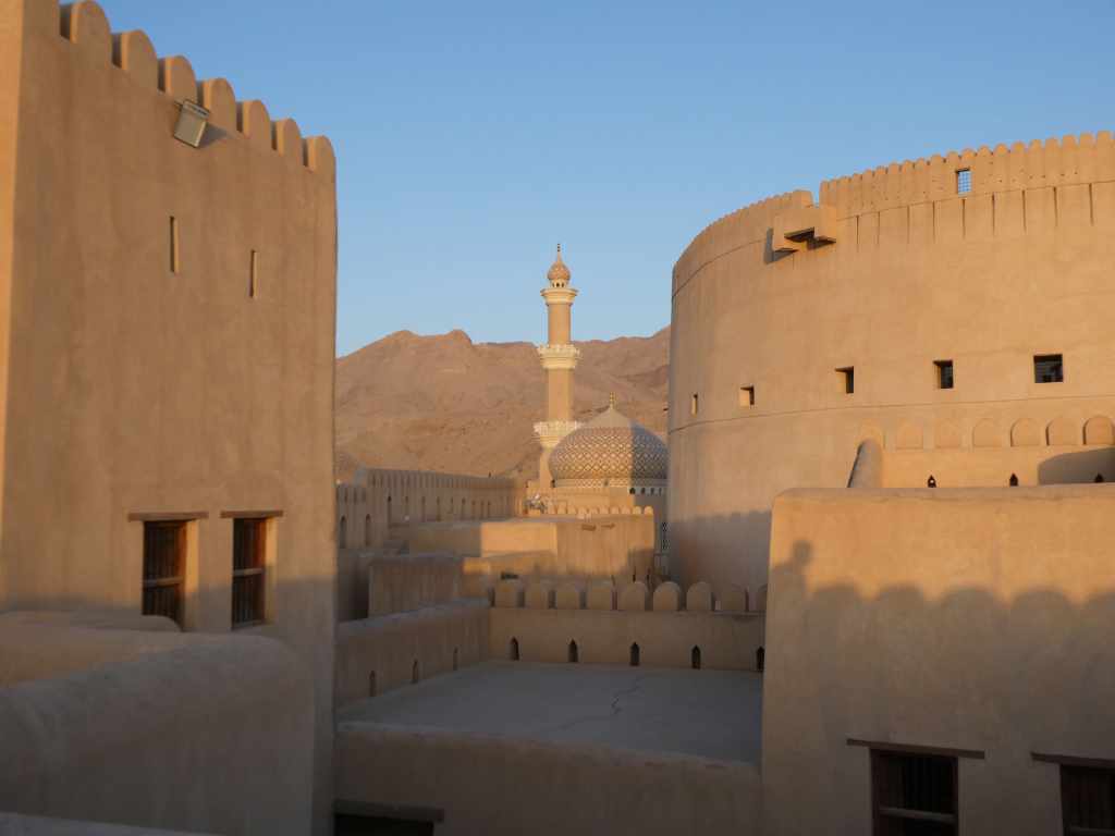 Vue panoramique d'un village traditionnel avec des murs en adobe et un minaret au fond, surplombé par des montagnes au coucher du soleil.