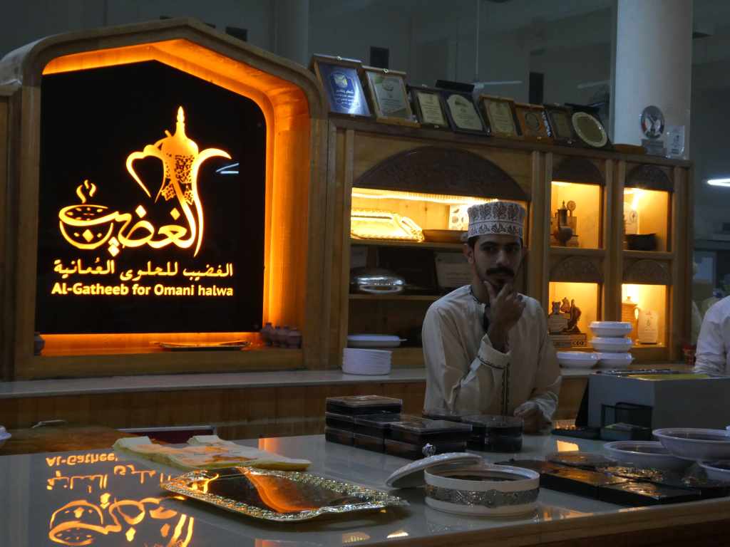 Un homme en tenue traditionnelle omanaise se tient derrière un comptoir avec des boîtes de halwa omanaise, devant un panneau lumineux affichant 'Al-Gatheeb for Omani halwa'.