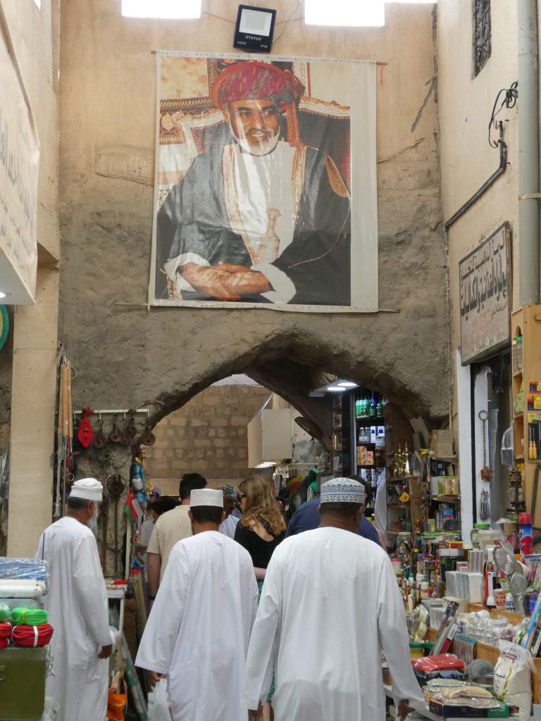 Une rue de marché animée avec des personnes portant des vêtements traditionnels, un grand portrait d'une figure royale suspendu au-dessus.